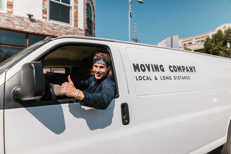 A male mover with long hair, wearing a black and white patterned headband and a dark blue shirt, is sitting inside the cab of a white van marked with the branding 'MOVING COMPANY LOCAL & LONG DISTANCE