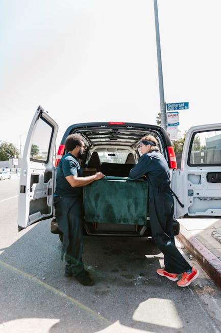A male mover with long hair, wearing a black and white patterned headband and a dark blue shirt, is sitting inside the cab of a white van marked with the branding 'MOVING COMPANY LOCAL & LONG DISTANCE