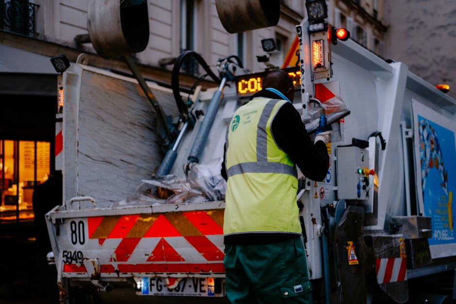 A worker from Removal Van Pimlico, dressed in a yellow high-visibility vest and dark clothing, operates control panels on a large waste collection vehicle parked outdoors during dusk. The vehicle's rear compartment is open, revealing a collection of cardboard boxes, plastic wrapping, and other recyclable materials, with some stacked and secured inside. The worker is facing the control panel, making adjustments or recording information as part of a home relocation or waste removal process. The scene is illuminated by the onboard lights of the waste truck, with a building visible in the background, indicating an urban environment. This image demonstrates the logistics involved in packing, loading, and waste management related to house removals or furniture transport services, with the removal van positioned on the pavement ready for disposal or transfer of goods. The overall setting emphasizes logistical operations in a city context, aligning with the services offered by Removal Van Pimlico for removals and waste clearance.