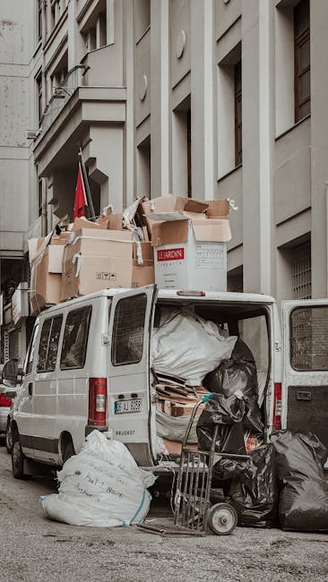 A white cargo van parked on a city street next to a multi-storey building with beige facades and rectangular windows. The van's rear doors are open, revealing a large amount of packed items, including black trash bags, cardboard boxes, and padding materials. On top of the van, several open and closed cardboard boxes are stacked, some with visible labels. A hand truck or dolly with additional boxes is positioned beside the van, indicating a loading process typical of home relocation or furniture transport. The scene is outdoors with natural daylight, capturing the logistics involved in house removals, with a focus on loading and packing materials as part of the moving service by Removal Van Pimlico.