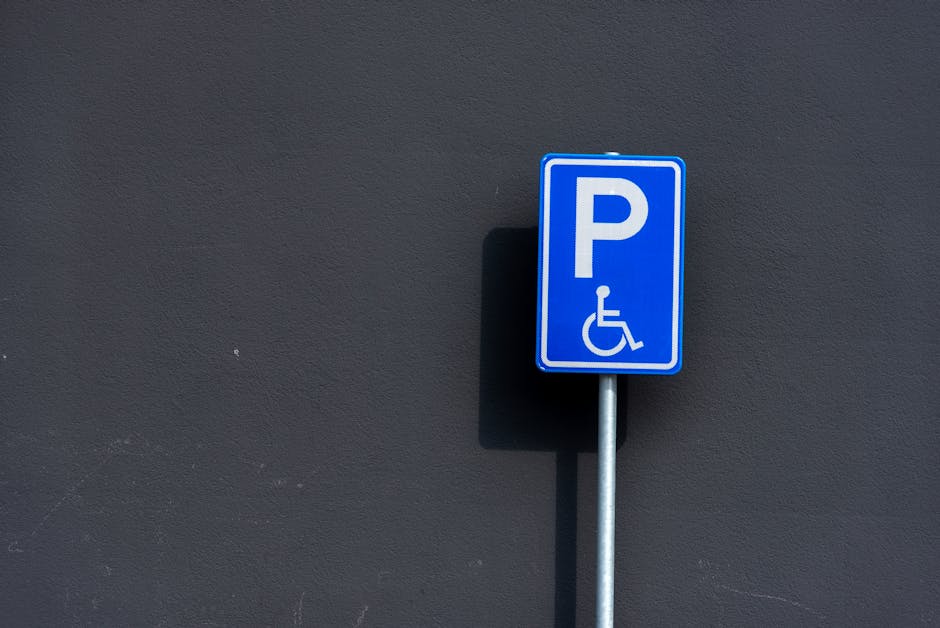 A vertical metal signpost displaying a reserved parking sign with a blue wheelchair symbol indicating accessibility, and a smaller sign below that reads 'VAN ACCESSIBLE'; the sign is positioned outdoo