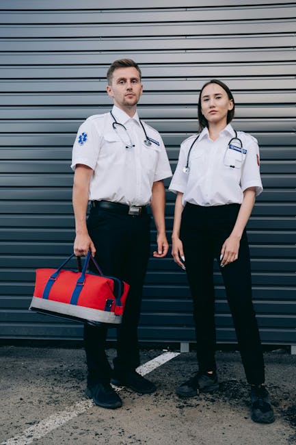 Two emergency medical professionals, a male and a female, standing outside a building with a dark grey, horizontal metal cladding door behind them. The male is holding a red and grey medical bag with 